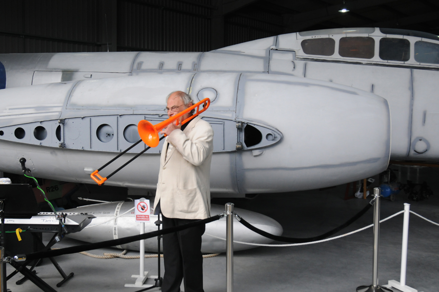 Tony Sheppard stands in front of an aircraft fuselage, playing an orange trombone. A keyboard is visible at the far left of the image.
