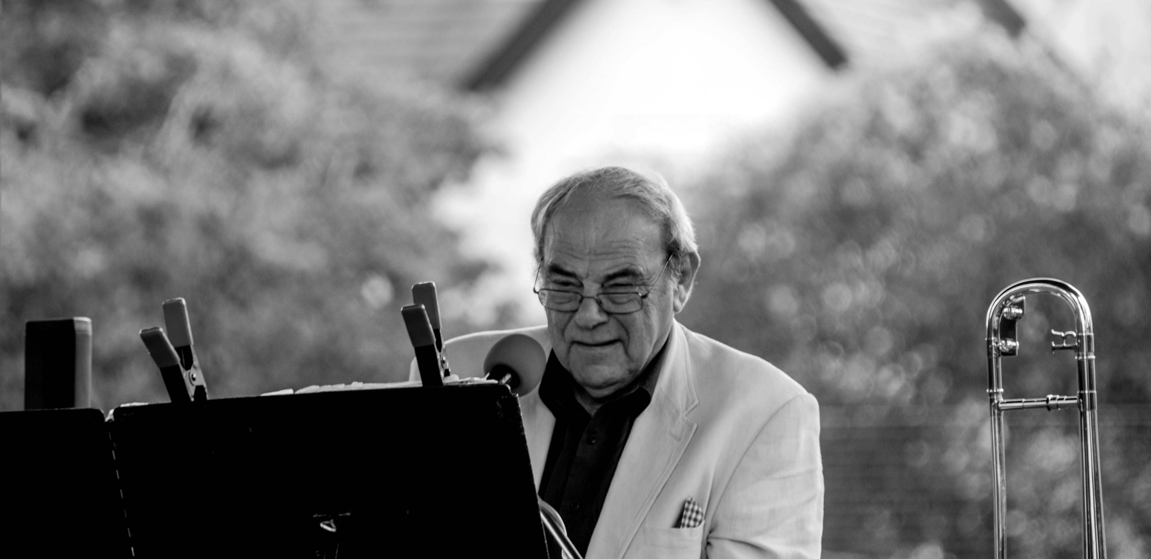 Tony Sheppard playing keyboard in an outdoor bandstand, with trees out of focus in the far distance. He wears a small smile of concentrated happiness. A trombone is visible by his left hand.