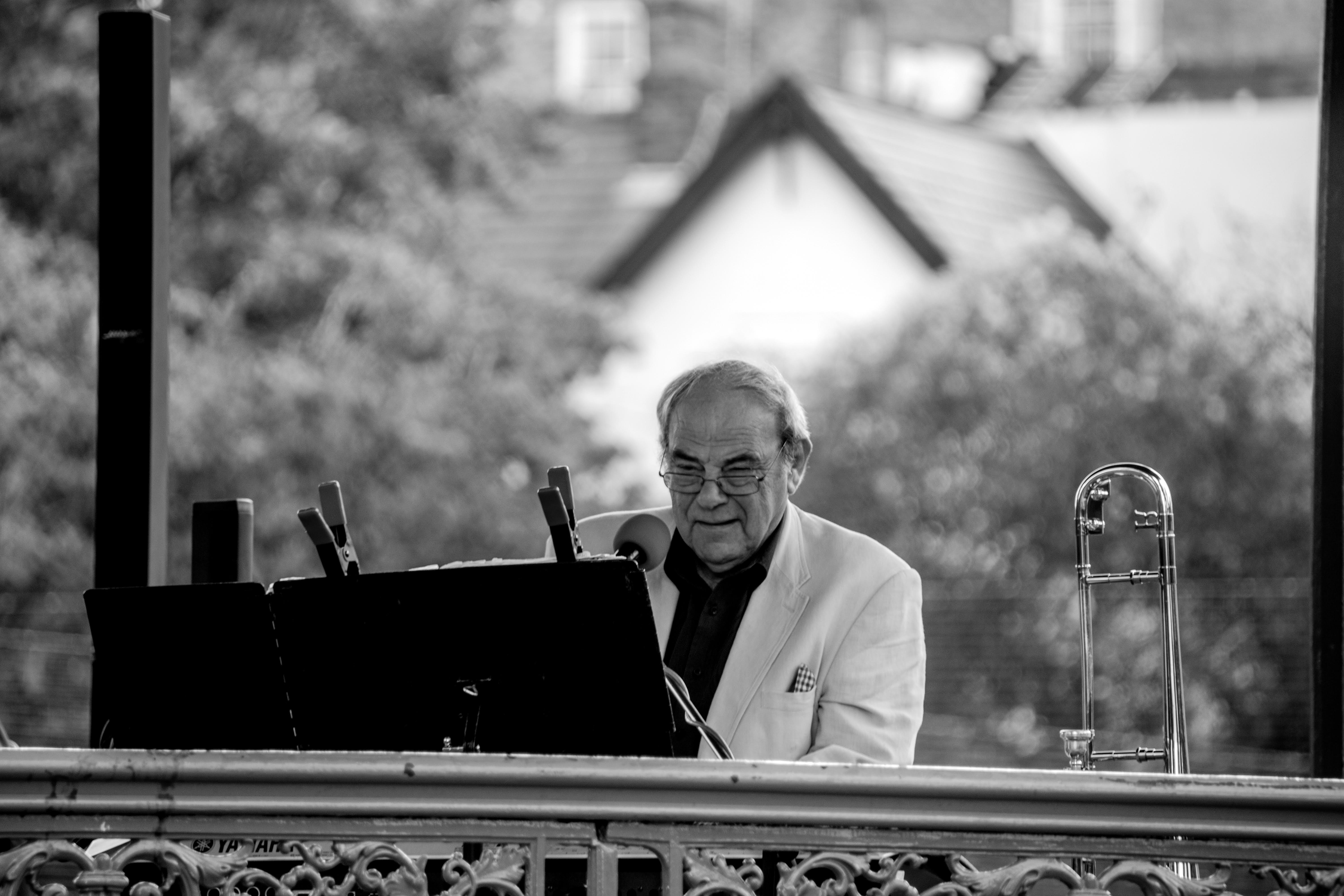 Tony Sheppard playing keyboard in an outdoor bandstand, with trees out of focus in the far distance. He wears a small smile of concentrated happiness. In front of him is a music stand, with music held down with what look like large clips from jumper cables. His trombone is on its stand by his left hand.