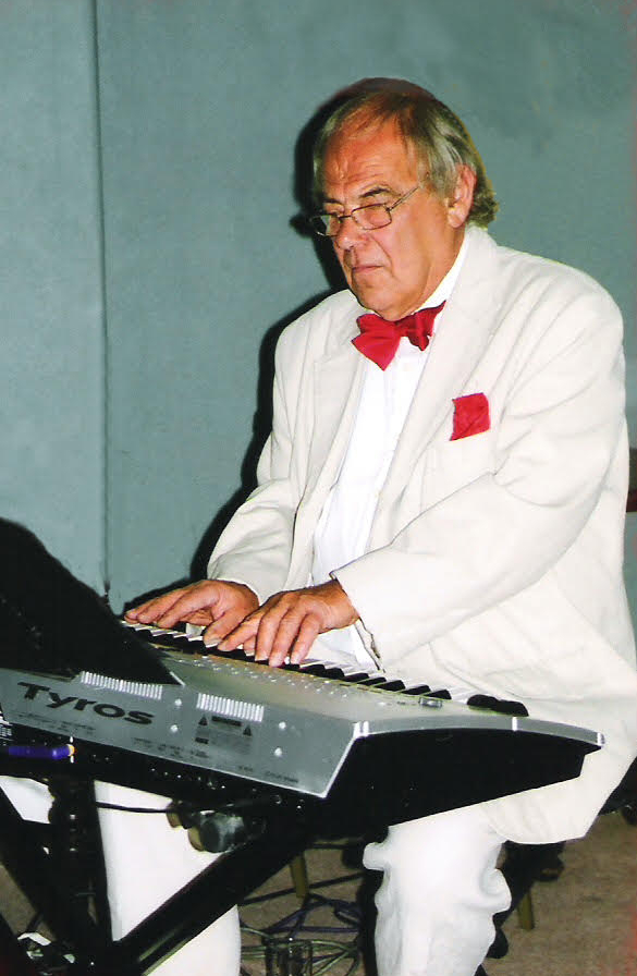 Tony Sheppard, seated in a white tuxedo with red bow tie playing a Tyros synthesizer. Tony's expression is serious or sad.